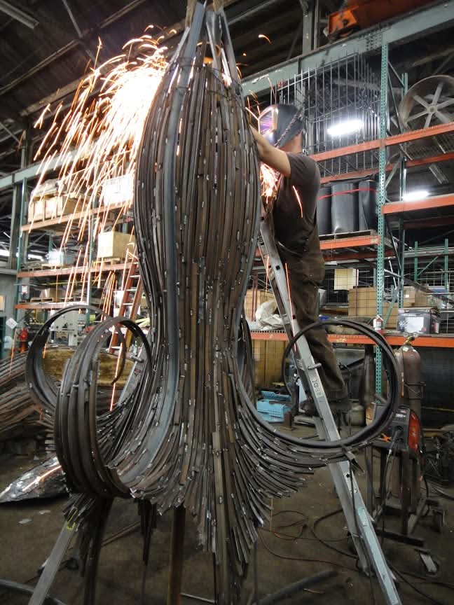 Large steel sculpture under fabrication, sparks flying as a worker grinds from a ladder