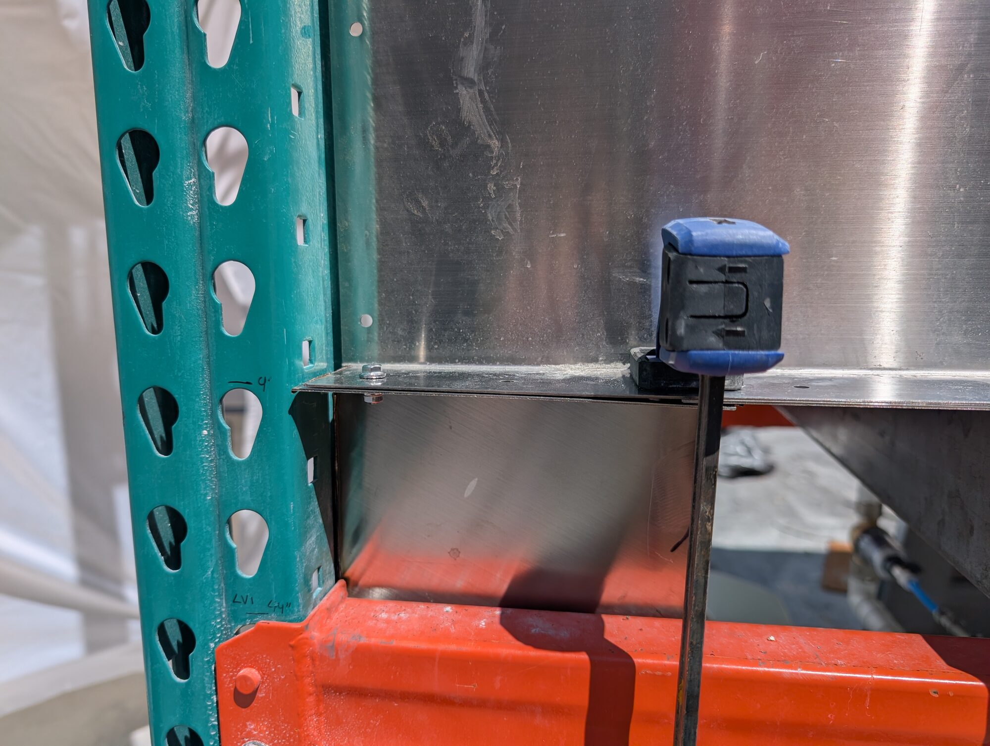 A tape measure clamped to the stainless hopper where it meets the teal racking upright, a 4 inch mark penciled on the steel