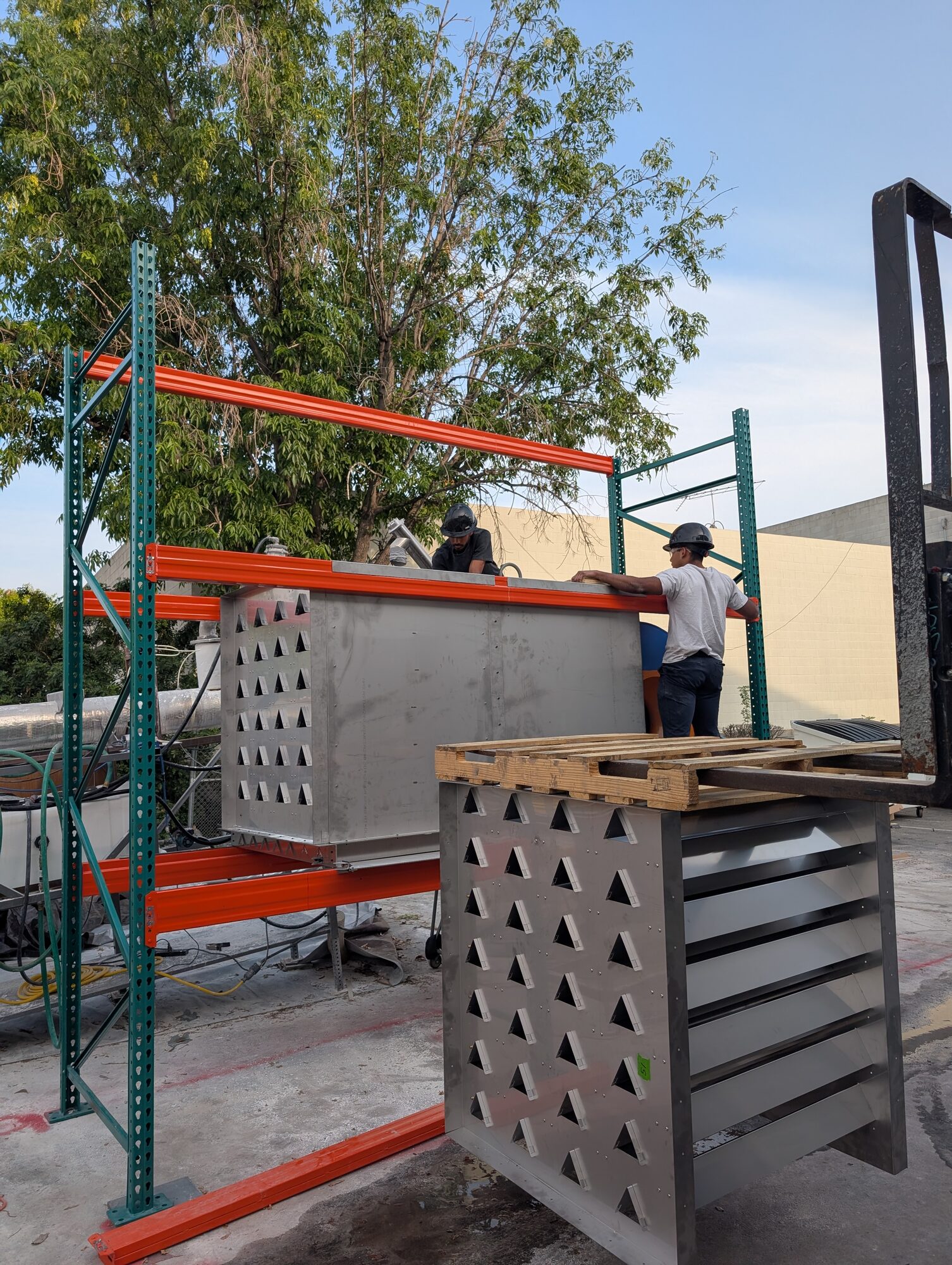 Two people in hard hats guiding a large stainless assembly onto orange pallet racking, a forklift mast at the right